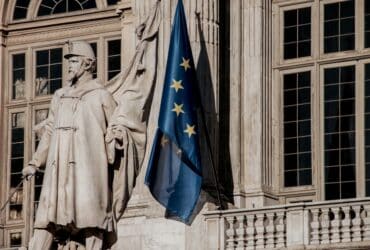 European Union flag waving beside a historic statue on stone building facade.