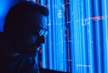 A man in sunglasses intently studies a vibrant blue holographic screen, symbolizing digital technology.