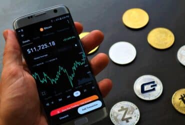 Close-up of a smartphone app showing Bitcoin trading details with crypto coins on a black surface.