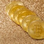 A stack of gold Bitcoin coins placed on a shimmering gold glitter background.