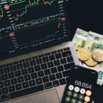 A laptop displaying stock charts with Bitcoin, Euros, and a cellphone calculator, showcasing financial analysis.