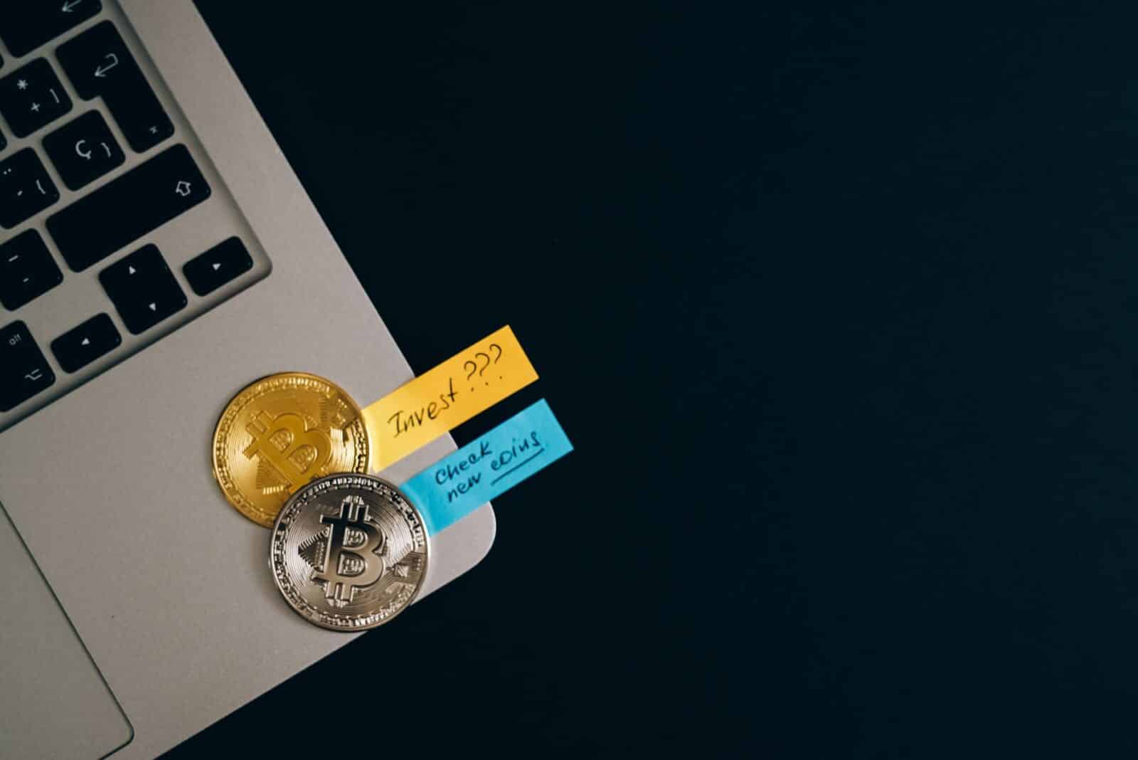Bitcoin coins placed on a laptop keyboard with sticky note reminders about investing and checking new coins.