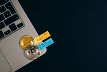 Bitcoin coins placed on a laptop keyboard with sticky note reminders about investing and checking new coins.