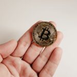 A close-up image of a Bitcoin coin held in a human hand against a white background.