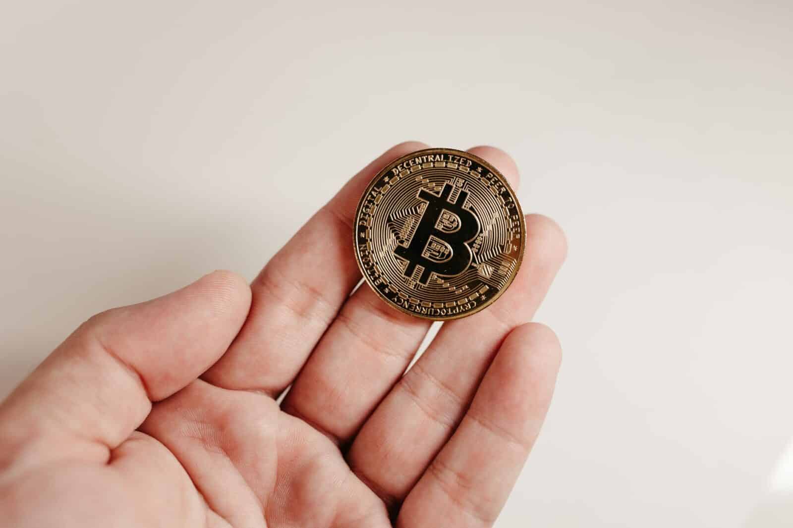 A close-up image of a Bitcoin coin held in a human hand against a white background.