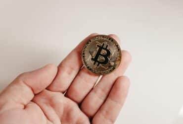 A close-up image of a Bitcoin coin held in a human hand against a white background.