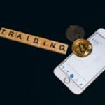 Cryptocurrency trading concept featuring Bitcoin, Scrabble tiles, and a smartphone on a black surface.