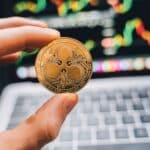 A close-up view of a hand holding a Ripple cryptocurrency coin with a blurred computer in the background.