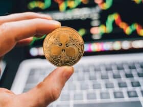A close-up view of a hand holding a Ripple cryptocurrency coin with a blurred computer in the background.