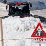 Snowplow clearing path on Swiss mountain, sign warns of avalanche danger.