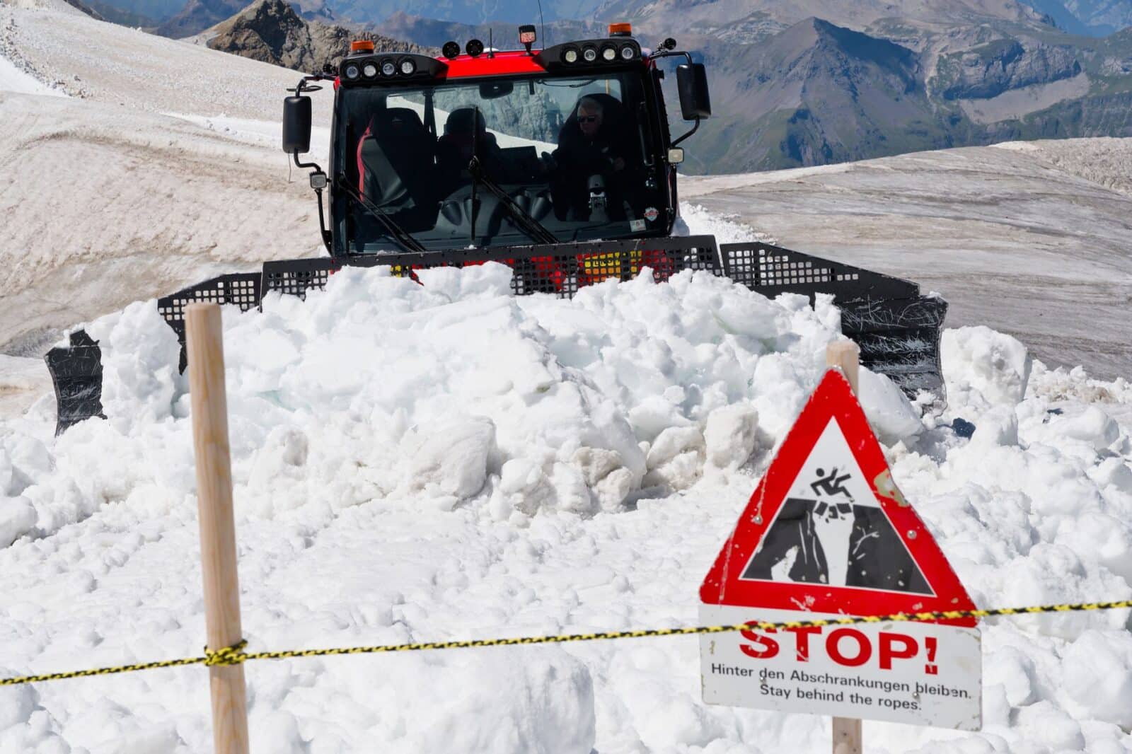 Snowplow clearing path on Swiss mountain, sign warns of avalanche danger.
