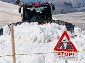 Snowplow clearing path on Swiss mountain, sign warns of avalanche danger.