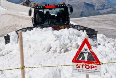 Snowplow clearing path on Swiss mountain, sign warns of avalanche danger.