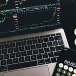 A laptop with trading charts, smartphone calculator, and bitcoin coins depicting cryptocurrency trading.