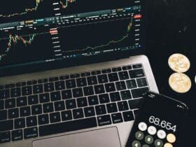 A laptop with trading charts, smartphone calculator, and bitcoin coins depicting cryptocurrency trading.