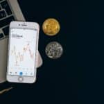 Smartphone displaying Bitcoin price chart alongside Bitcoin and Ethereum coins on black background.