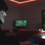 A person wearing a hacker mask operates a computer in a dimly lit room with digital displays.