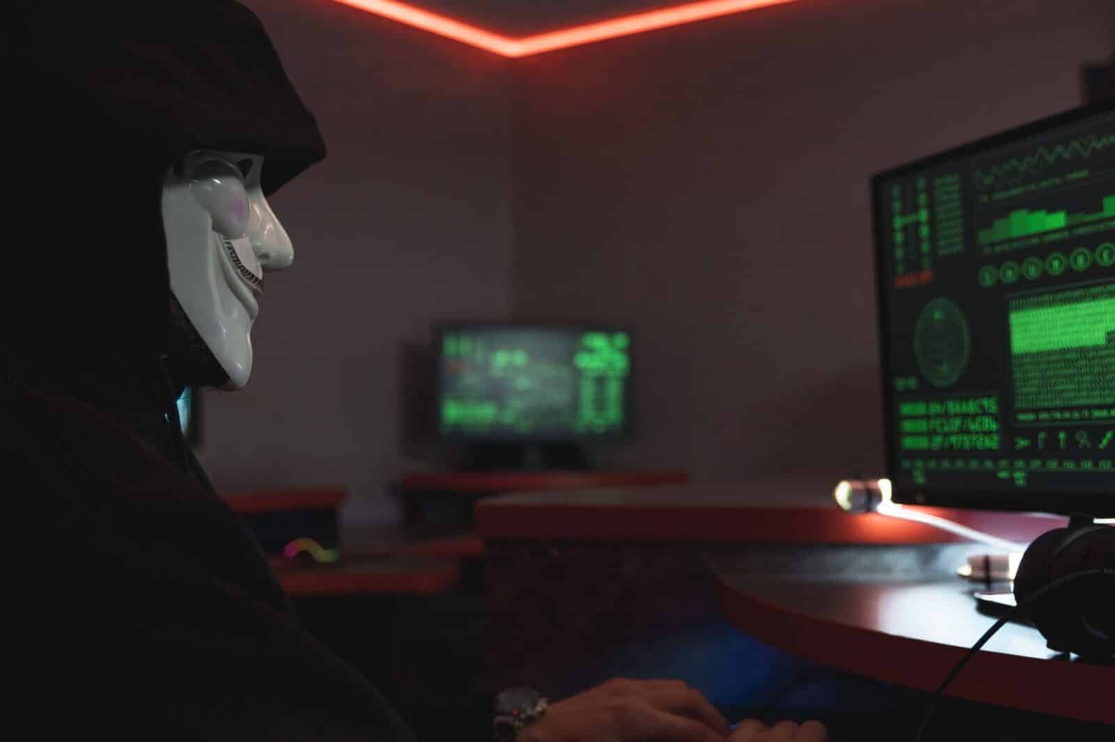 A person wearing a hacker mask operates a computer in a dimly lit room with digital displays.