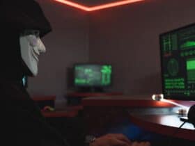 A person wearing a hacker mask operates a computer in a dimly lit room with digital displays.