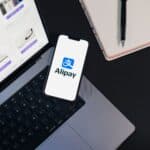 Smartphone displaying Alipay app on open laptop with online shopping site.