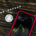 Smartphone showing bitcoin price with a physical coin and text representing cryptocurrency investment.