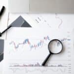Flat lay of stock market analysis documents with magnifying glass, pens, and glasses.