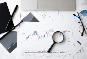 Flat lay of stock market analysis documents with magnifying glass, pens, and glasses.