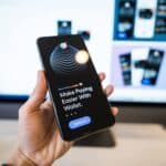 Hand holding smartphone displaying digital wallet app interface, blurred monitor in background.