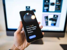 Hand holding smartphone displaying digital wallet app interface, blurred monitor in background.