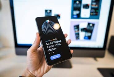 Hand holding smartphone displaying digital wallet app interface, blurred monitor in background.