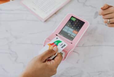 Hand using a contactless card on a pink POS terminal for a wireless transaction.