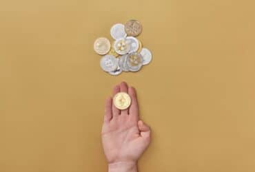 Close-up of a hand holding a cryptocurrency coin on a yellow background with scattered coins.